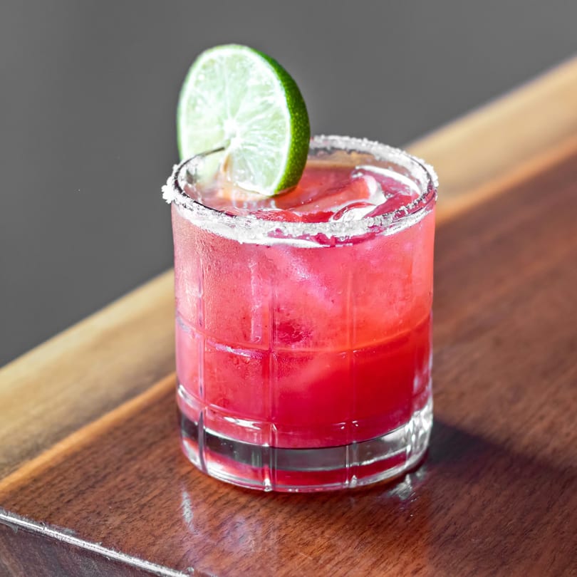 A vibrant red cocktail with ice, a salted rim, and a lime wedge on a wooden surface.