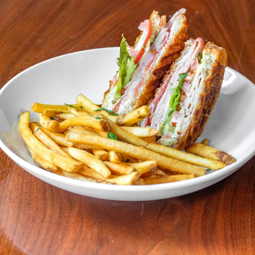 Club sandwich with crispy fries on a white plate, showcasing layers of meat, lettuce, and tomato.
