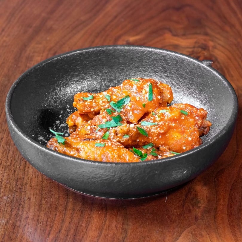 Spicy chicken wings coated in orange sauce, sprinkled with cheese and parsley, in a black bowl.