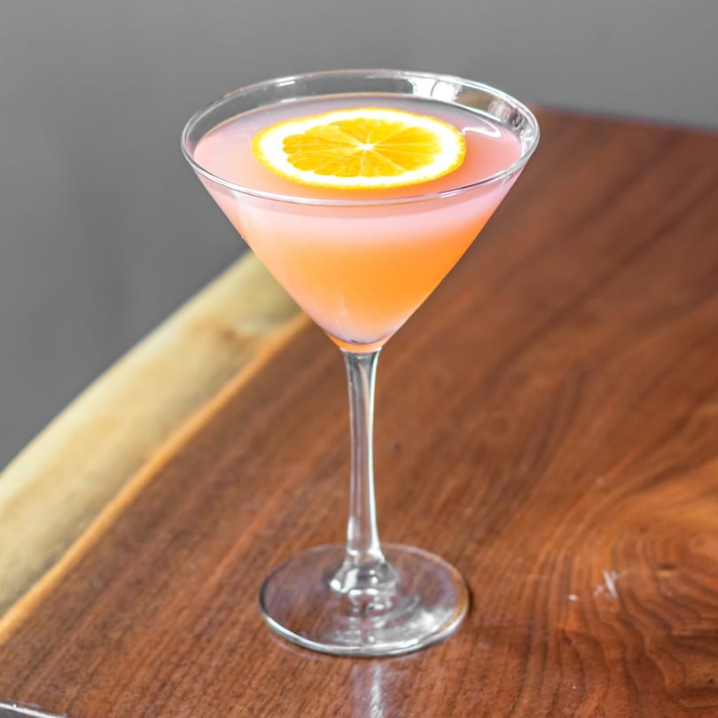 Pink cocktail in a martini glass, garnished with an orange slice, on a wooden table.