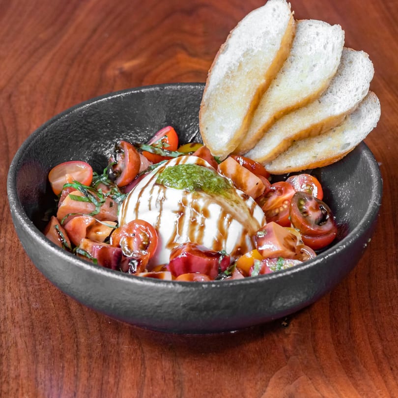 Burrata cheese with cherry tomatoes, pesto, balsamic glaze, and toasted bread slices in a black bowl.