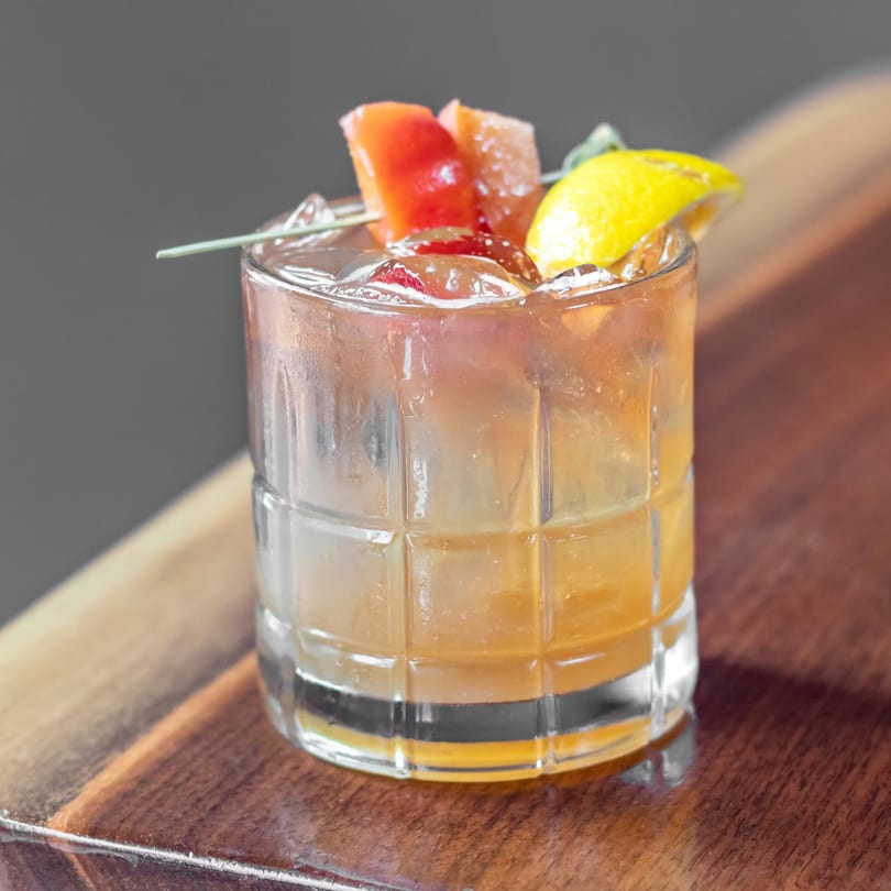 A refreshing cocktail with ice, garnished with grapefruit, cherries, and a lemon wedge on a wooden surface.